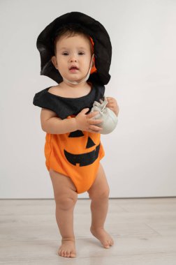 Beyaz arka planda kafatası tutan Cadılar Bayramı için balkabağı kostümü giymiş şirin beyaz çocuk.