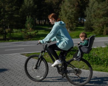 Küçük oğluyla bisiklete binen beyaz bir kadın arkasında çocuk koltuğunda oturuyor.