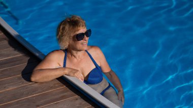 Güneş gözlüklü yaşlı bir kadın havuzda yüzüyor. Emeklilikte tatil