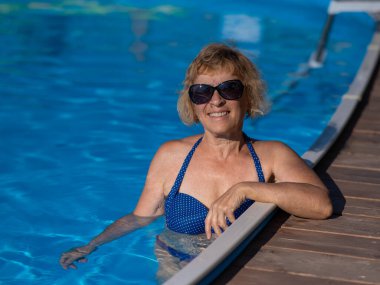 Güneş gözlüklü yaşlı bir kadın havuzda yüzüyor. Emeklilikte tatil