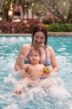 Beyaz bir kadın tatilde havuzda yüzerken oğlunu kucağına alıyor. Annesi küçük çocuğa yüzmeyi öğretiyor.