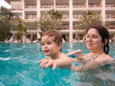 Beyaz bir kadın tatilde havuzda yüzerken oğlunu kucağına alıyor. Annesi küçük çocuğa yüzmeyi öğretiyor.