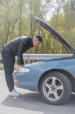 Beyaz adam arabasının kaputunun altına bakıyor.