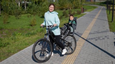 Beyaz kadın bisiklete biniyor. Yanında da oğlu var. Parkta çocuk koltuğunda oturuyor.