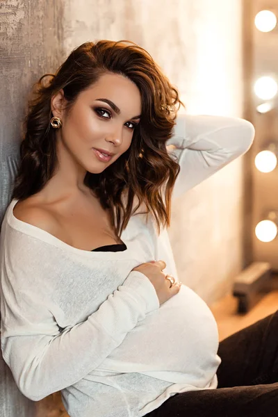 Femme enceinte européenne en chemisier blanc et pantalon noir est assis près du mur dans une femme enceinte sombre plat, élégant et heureux dans les appartements, belle femme enceinte avec les cheveux foncés — Photo