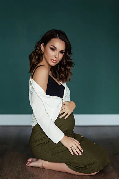 Pregnant european woman sits on the floor in white blouse and green skirt with beige hat in dark flat, stylish and happy pregnant woman in apartments, beautiful pregnant woman in dark studio