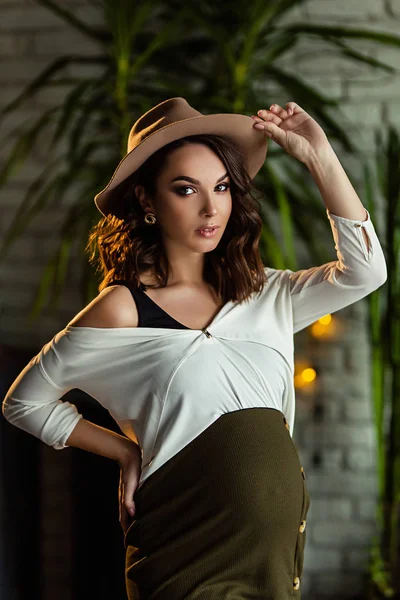 Pregnant european woman in white blouse and green skirt with beige hat in dark flat looks to the camera, stylish and happy pregnant woman in apartments, beautiful pregnant woman in dark studio