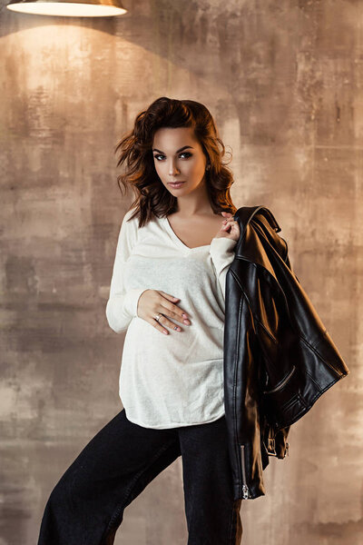 Pregnant european woman in white blouse and black pants in dark flat, stylish and happy pregnant woman in apartments, beautiful pregnant woman with dark hair