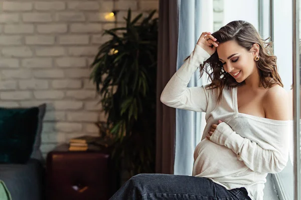 Pregnant european woman in white blouse and black pants is sitting on the windosill, stylish and happy pregnant woman in apartments, beautiful pregnant woman with dark hair