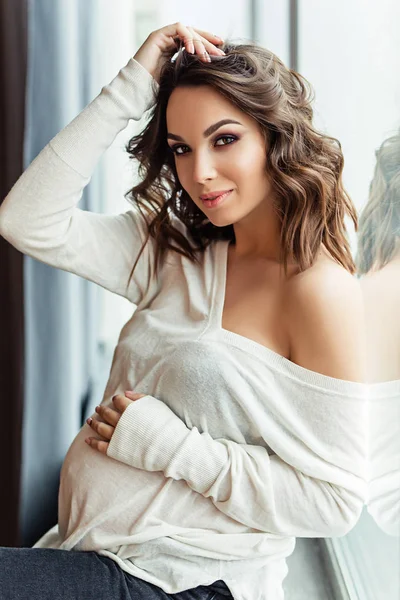 Pregnant european woman in white blouse and black pants is sitting on the windosill, stylish and happy pregnant woman in apartments, beautiful pregnant woman with dark hair