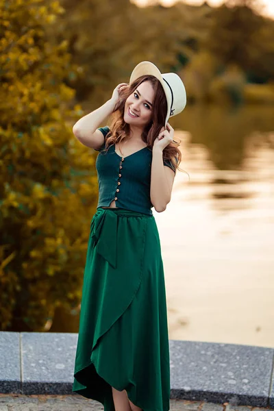 Young european woman is standing near the lake, beautiful european woman in nature, pretty young woman in hat and green dress near the river