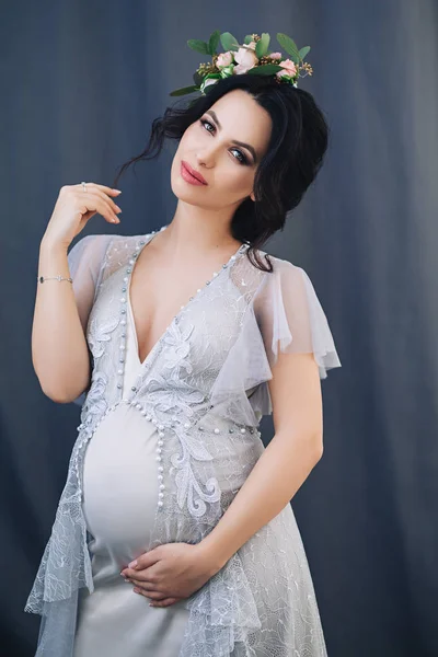 Femme enceinte européenne sur fond gris, jeune femme européenne en attente d'un enfant, femme prenant aux cheveux noirs en robe longue et légère dans un appartement blanc, belle jolie future mère avec couronne — Photo