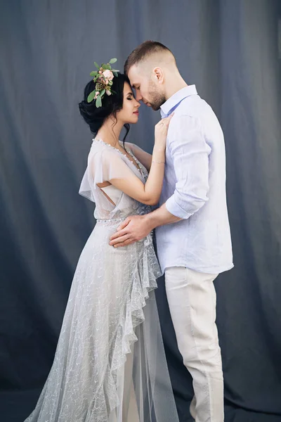 Pregnant european woman with her husband on gray background, young european couple waiting for a child, prenant woman with black hair in long light dress with wreath of flowers, happy european future