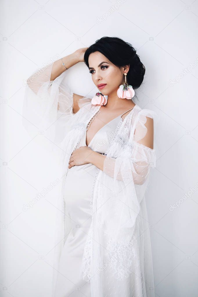Pregnant european woman in white apartment, young european woman waiting for a child, prenant woman with black hair in long light dress in white apartment, beautiful pretty future mother with stylish