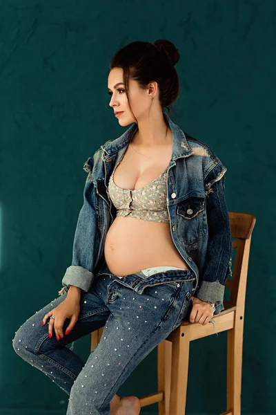 Pregnant european woman on blue background, beautiful young european woman waiting for a child, prenant woman with dark hair in jeans is sitting on a chair