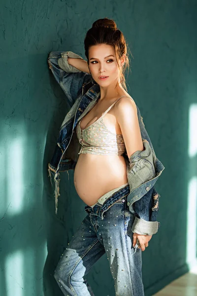 Pregnant european woman on blue background, beautiful young european woman waiting for a child, prenant woman with dark hair in jeans is girl looking away