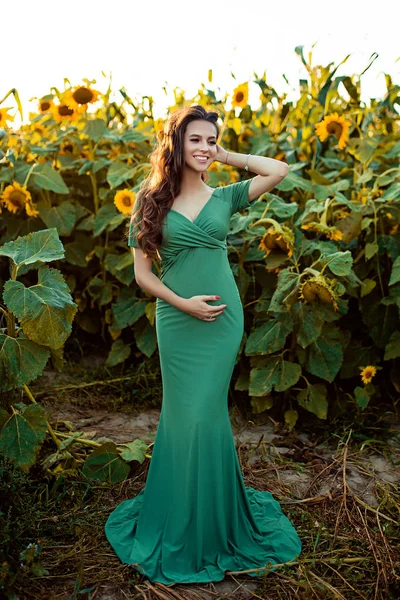 Pregnant european woman in a field of sunflowers, beautiful young european woman waiting for a child, prenant woman with dark hair in long green dress is smiling on the nature