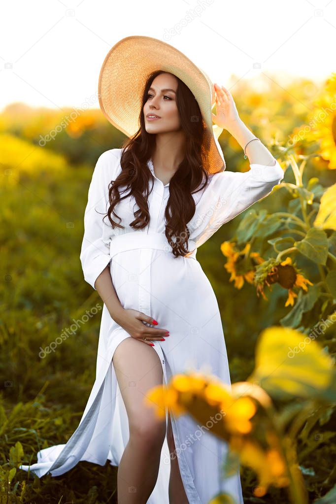 Pregnant european woman in a field of sunflowers, beautiful young european woman waiting for a child, prenant woman with dark hair in white long dress with hat on the nature