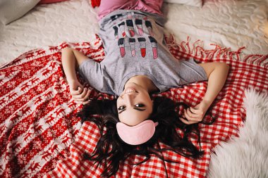 Genç Avrupalı bir kadının Noel atmosferindeki portresi, uzun siyah saçlı ve parlak makyajlı beyaz pijamalı güzel bir kadın, uyku maskesiyle yatağa uzanır.