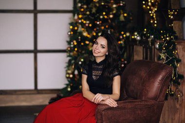 Karanlık stüdyoda, Noel ağacıyla birlikte genç Avrupalı bir kadının portresi, siyah bluzlu, uzun siyah saçlı ve parlak makyajlı beyaz bir kadın, koltukta oturuyor.