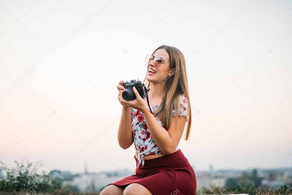 Attractive tourist girl with brown hair wearing hat, sunglasses, holding camera and smiling, traveling, portrait.