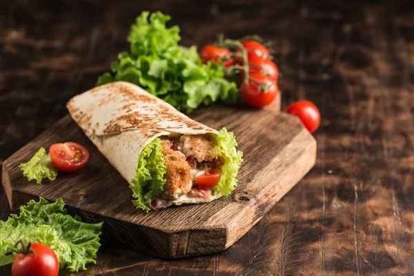 Concept of street fast food - Roll with chicken and fresh cherry tomatoes and lettuce on a wooden Board on a dark background. Close-up with copy space. Horizontal orientation.