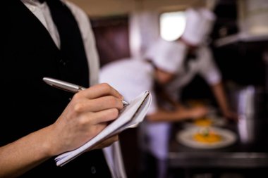 Mid section of female waitress noting an order on notepad in kitchen clipart