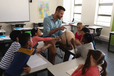 Male teacher teaching his kids about geography by using globe in classroom of elementary school clipart