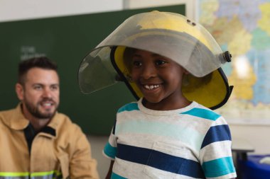 Front view of a happy black schoolboy wearing fire helmet and a smiling firefight behind him in classroom of elementary school clipart