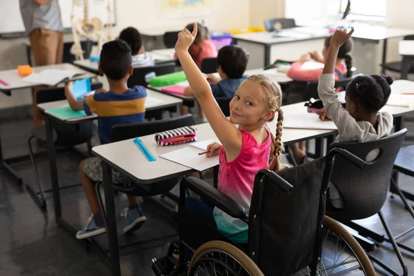 Smiling disable schoolgirl looking at camera and raising hand in classroom of elementary school