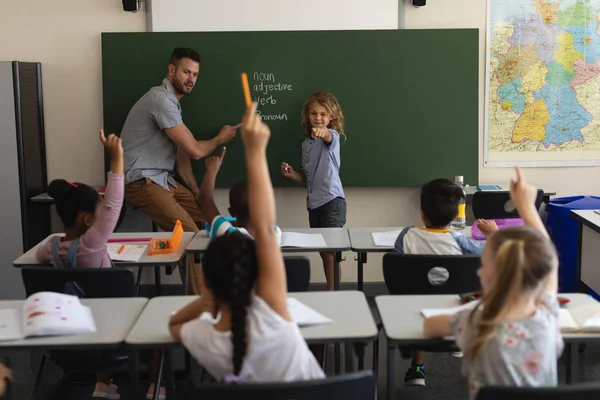 Rear view of schoolboy pointing finger towards classmates in classroom of elementary school