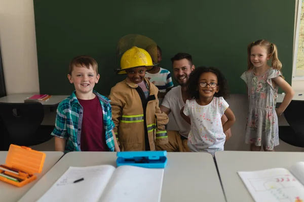 Front view happy male firefighter with schoolkids looking at camera and one of them has got firefight uniform in classroom of elementary school