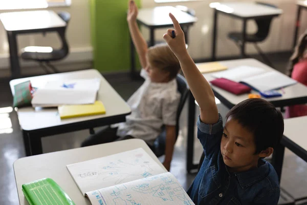 坐在桌旁的小学生在教室里举手的高视角 — 图库照片