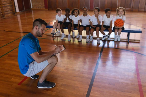 Basketball coach writing on clipboard and schoolkids sitting on bench at basketball court in school