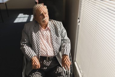 Front view of disable senior man sitting on wheelchair and looking outside through window at home clipart
