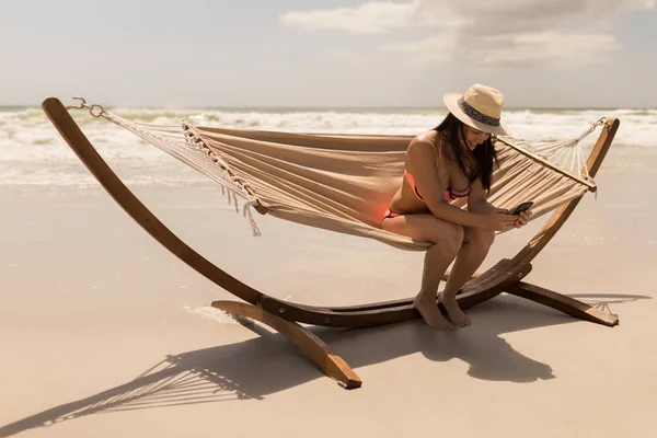 在阳光下坐在沙滩上的吊床上 戴着帽子戴着手机的美丽快乐年轻女子的正面景色 — 图库照片