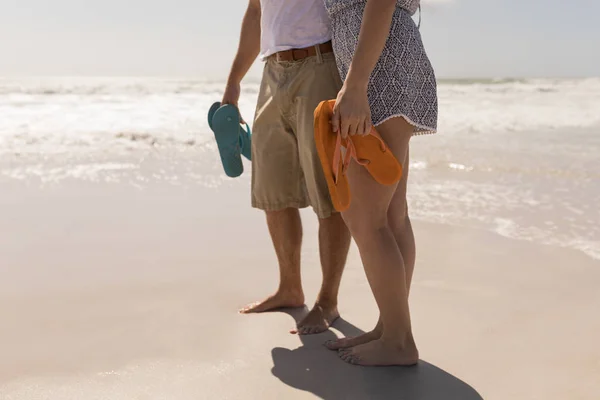 在阳光下在沙滩上拿着鞋子的年轻浪漫夫妇的低部分 — 图库照片
