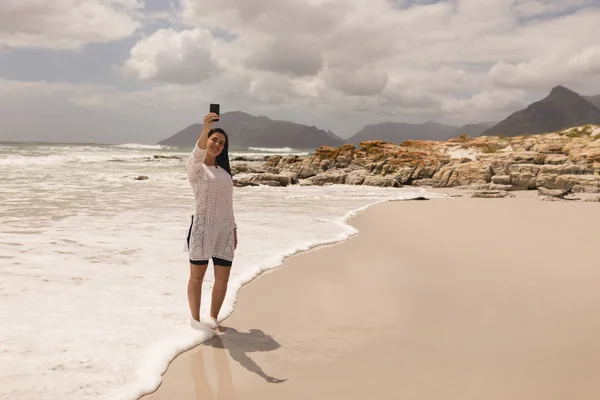 年轻女子在阳光下拿着手机在海滩上自拍的正面看法 — 图库照片