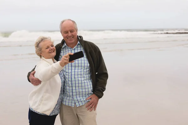 在背景中 活跃的高级夫妇带着手机站在海滩上的侧视图 — 图库照片