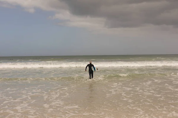 シニアの女性サーファーがサーフボードとビーチの上を歩いての背面図 — ストック写真
