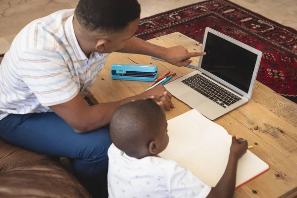 非洲裔美国人的父亲在舒适的家里用笔记本电脑在桌子上帮儿子做作业的高角度 — 图库照片