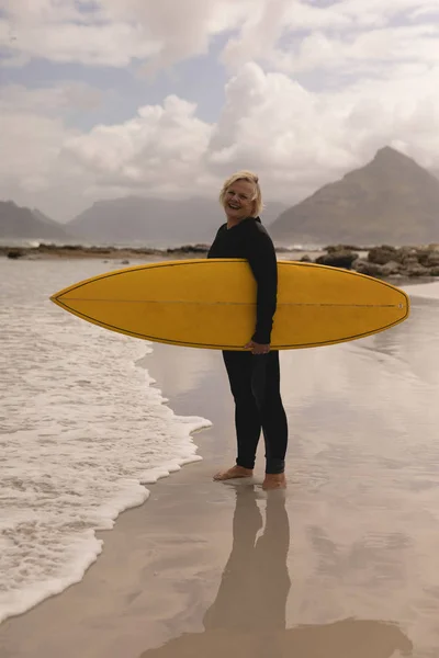 ビーチにサーフボードを抱えて立っているアクティブ シニア女性の側面図 — ストック写真