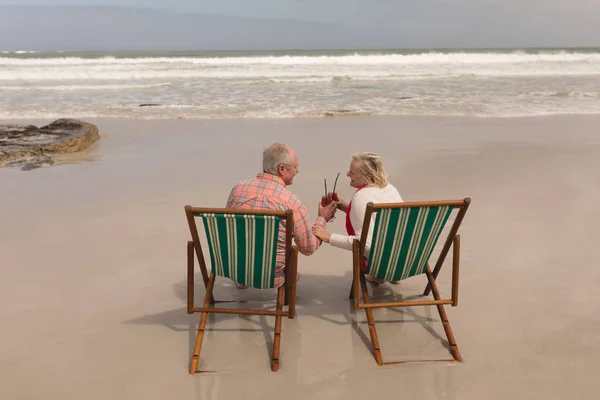 在以海洋为背景的海滩上 在阳光躺椅上品尝鸡尾酒饮料的活跃的资深夫妇的背影 — 图库照片