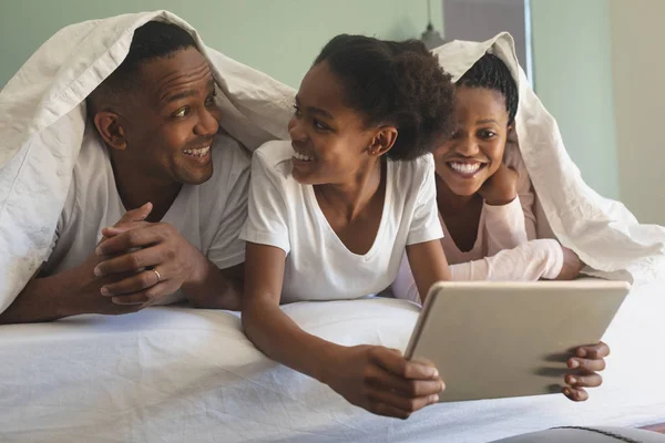 デジタル タブレットを使用して 自宅の寝室のベッドの上 毛布の下で幸せなアフリカ系アメリカ人の家族の正面図 — ストック写真