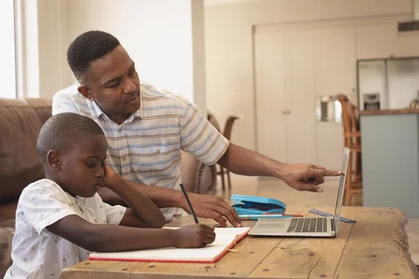 非洲裔美国人父亲在舒适的家里用笔记本电脑在桌子上帮儿子做作业的侧视图 — 图库照片