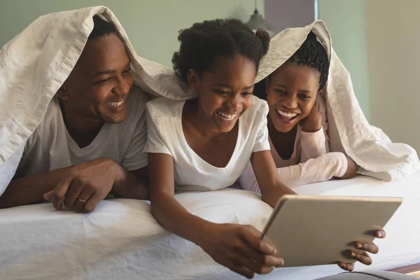デジタル タブレットを使用して 自宅の寝室のベッドの上 毛布の下で幸せなアフリカ系アメリカ人の家族の正面図 — ストック写真