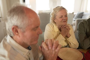 Side view of senior couple praying together before having meal on dining table at home clipart