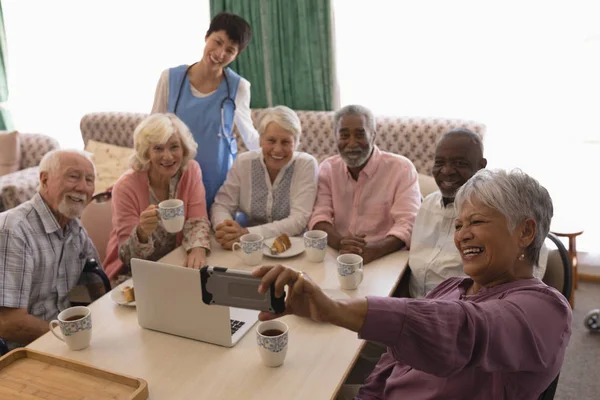 老年妇女拿着手机在一张桌子周围自拍的正面看法 女医生站在养老院 — 图库照片