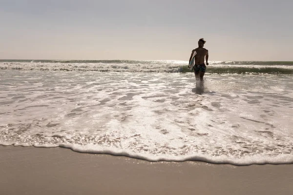 太陽の光のビーチを走るサーフボードと上半身裸の若い男性サーファーの正面図 — ストック写真