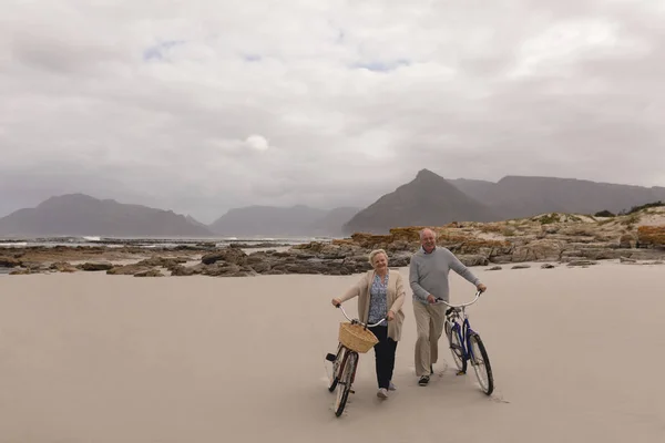 活跃的高级夫妇步行与自行车在海滩与山的背景的前面的看法 — 图库照片